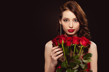 Attractive young woman holding bouquet of red roses and looking at camera isolated on black