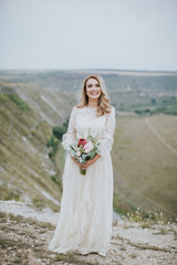 Fototapeta na wymiar Smiling young blonde caucasian bride in a white, modern lightweight dress posing. Wedding bouquet in her hands. Happy vintage hipster bride, warm colors.