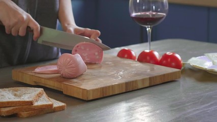 Woman cuts club sandwich in a half by sharp knife on a wooden board at the kitchen, making of the fastfood at home. Close-up. Girl's hands chop ham or sausage for a sandwich, breakfast. - Powered by Adobe
