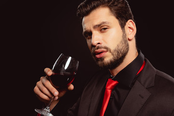 Handsome man in formal wear and red tie looking at camera while holding glass of red wine isolated on black