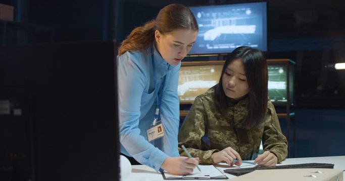 Caucasian young woman showing something to Asian female soldier and educating her on army strategy in controlling office. Educational military center for women concept. Soldier and official discussion