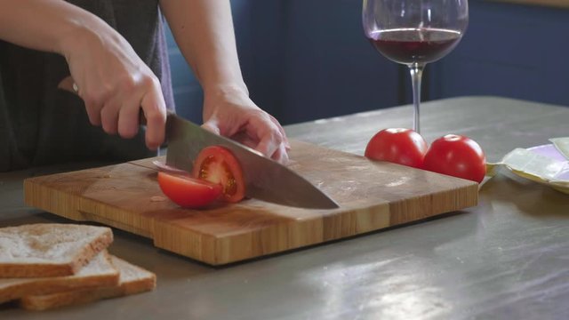 Woman cuts club sandwich in a half by sharp knife on a wooden board at the kitchen, making of the fastfood at home. Close-up. Girl's hands chopped tomatoes for a sandwich, breakfast