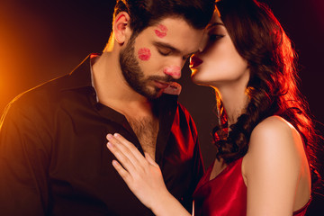 Elegant woman kissing handsome boyfriend with lipstick prints of face on black background with lighting