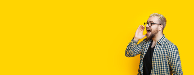 Blonde caucasian man with beard and eyeglasses is screaming on a yellow background with free space