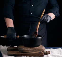 male cook in black uniform and latex gloves stirs a vintage wooden spoon food in a cast-iron pan with a handle