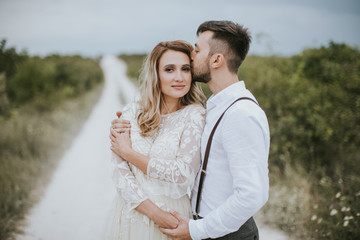 Smiling bride and groom spending time together. Posing on the mountain hills background. Dressed in white dress beautiful blonde caucasian bride and handsome groom. Hugs, kissess and enjoy the company