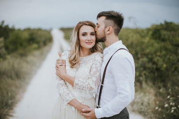 Obraz premium Smiling bride and groom spending time together. Posing on the mountain hills background. Dressed in white dress beautiful blonde caucasian bride and handsome groom. Hugs, kissess and enjoy the company