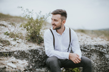 Handsome smiling groom dressed in white shirt and gray pants posing on a beautiful mountain hills landscape