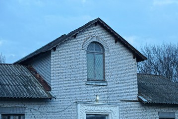 the attic of a white brick house with one large window under a gray slate roof against the sky in the evening street