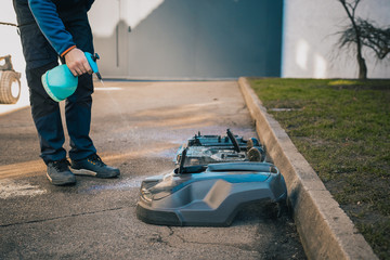 Man cleaning the underside of a robotic autonomous lawnmower with the help of a cleaning solvent as part of a service plan. Gunk will be flying out.