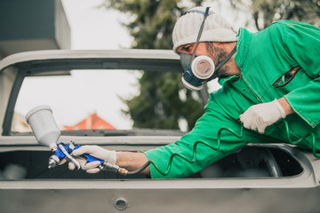 Caucasian man is spraying color with a compressed air paint gun on the vintage car as a restoration...