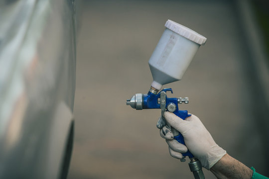 Caucasian man is spraying color with a compressed air paint gun on the vintage car as a restoration project. Man wearing protective equipment such as mask and gloves, professional automotive painter.