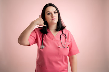 Beautiful woman doctor with stethoscope, wearing pink scrubs showing call me gesture