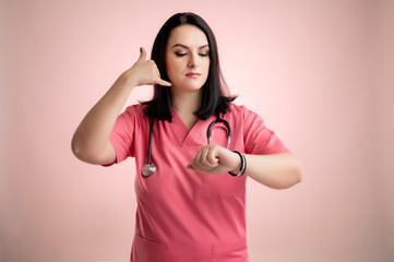 Beautiful woman doctor with stethoscope, wearing pink scrubs showing call me gesture and looking...