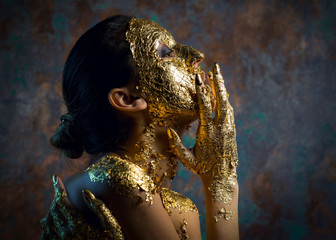 Girl with a mask on her face made of gold leaf. Gloomy studio portrait of a brunette on an abstract background.