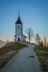 Fototapeta na wymiar Path towards cute fantastic charming Saint Primoz church on a small hill with mountains in background at sunset, Jamnik village, Slovenia, Europe with picturesque church in early spring.