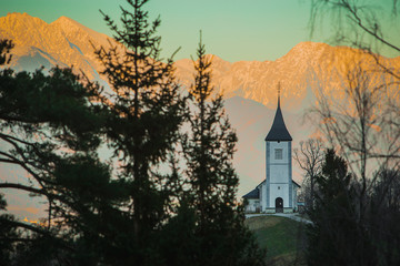 Fototapeta premium Fantastic charming Saint Primoz church on a small hill with colorful mountain backdrop in background at sunset, Jamnik village, Slovenia, Europe with picturesque church in early spring.