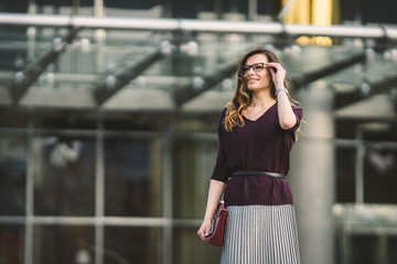 Business woman standing on street against office building. City business woman working. Portrait business woman smiling with glasses. Woman posing outside office building summer business outfit