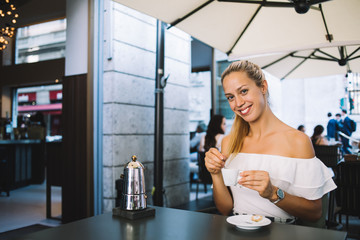 Fototapeta na wymiar Portrait of smiling caucasian blonde 20s woman enjoying aroma coffee on breakfast in cafeteria recreating on vacation holidays, cheerful young hipster girl looking at camera holding espresso cup