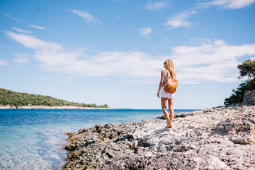 Young woman tourist in casual wear and with rucksack standing on rocky shore near ocean looking at beautiful nature marine landscape, back view of female traveler enjoying cal harbour in summer