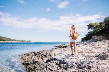 Blonde female caucasian explorer with backpack enjoying beautiful seashore having summer vacations,20s back view of woman traveler with rucksack looking at sky and calm nature near ocean