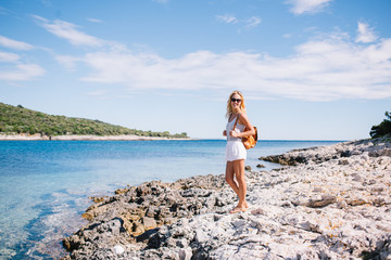 Portrait of cheerful caucasian female explorer with backpack satisfied with journey and holiday getting to ocean destination, smiling blonde woman 20s in sunglasses standing near seascape enjoying