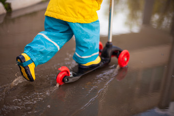 spring walks in any weather. the child rides a scooter through puddles in rubber boots and a waterproof suit, splashing water in all directions. Happy childhood with fun walks