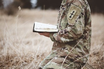 Shallow focus shot of a young soldier kneeling while holding an open bible in a field