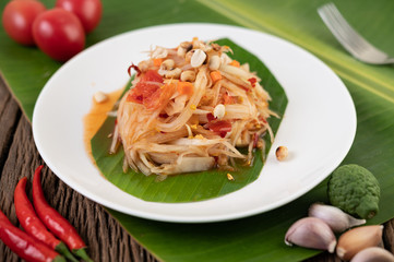 Thai papaya salad in a white plate on banana leaves with lime, tomatoes, eggplant, chili, garlic, peppers, salad, and peanuts.