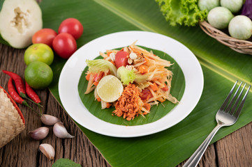 Thai papaya salad in a white plate on banana leaves with lime, tomatoes, eggplant, chili, garlic, peppers, salad, and peanuts.