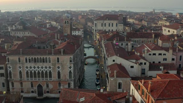 Aerial, tracking, drone shot overlooking a small channel and bridges, in middle of Venetian buildings, streets lights on, evening dusk, in Venezia, Italy