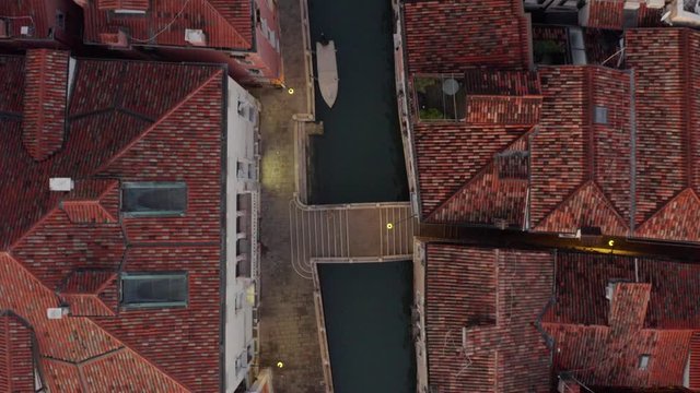 Aerial, top down, drone shot over a small bridge, at a small channel, in middle of Venetian buildings, streets lights on, evening dusk, in Venezia, Italy