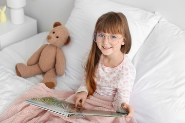 Cute little girl wearing glasses while reading book in bed