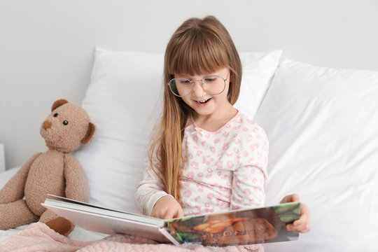 Cute little girl wearing glasses while reading book in bed