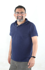 A smiling Indian man with a goatee beard wearing spectacles against a white background. looking to...