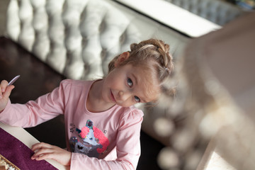 Beautiful little girl in a pink dress sits on a white sofa in a cafe, sunny day and beautiful vintage purple interior