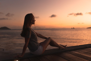 Fototapeta premium Beautiful girl at sunset on the sea in Thailand. Bright orange and yellow pink sky. Girl traveler enjoys the view. Woman in the tropics
