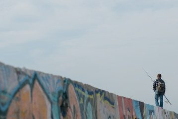 adult man with fishing rod and backpack standing on wall with graffiti