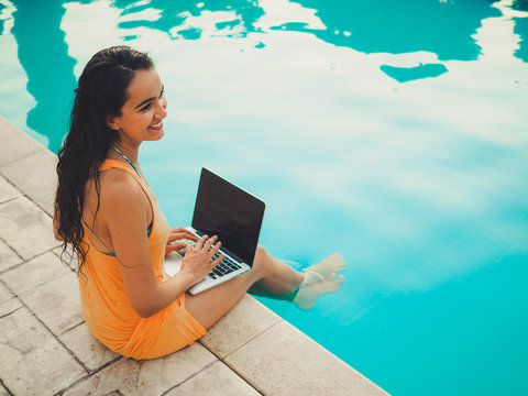 smiling happy remote online working digital nomad women with long hair and laptop sitting at a sunny blue water pool