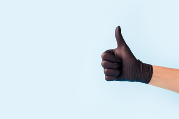 Closeup of female hand in black cosmetic medical hygiene pharmacy gloves showing thumbs up sign against pastel blue background, copy space, minimal concept