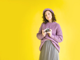 young girl in beautiful lilac clothes with a retro camera in hands on a yellow background