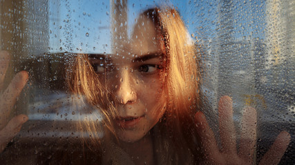The girl looks suspiciously behind the glass. There are raindrops on the glass. The girl has blond...