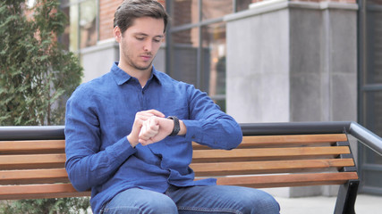 Fototapeta premium Young Man Using Smartwatch, Outdoor Sitting on bench