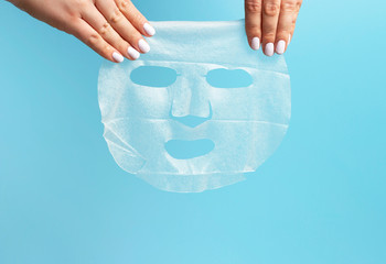 Female hands with white manicure hold fabric mask on a blue background.