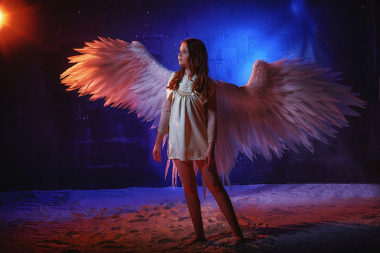 White angel on a dark background with colored lighting. The concept of war between good and evil. Girl with angel wings during a photo shoot with flour and loose powder