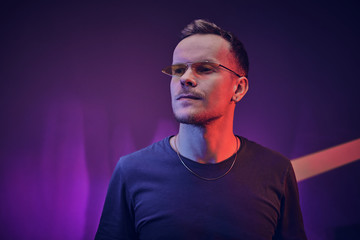Obraz premium Still and confident, male model posing in a neon studio for the photoshoot wearing casual clothes and yellow sunglasses, simple style looking bold and cool, slightly smirking