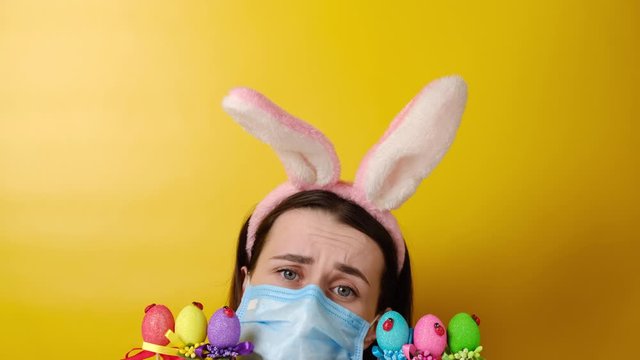Displeased young woman with virus mask, wears fluffy ears, sad to celebrate Easter alone, holds colored eggs on yellow background with copy space. Coronavirus outbreak Holiday concept.