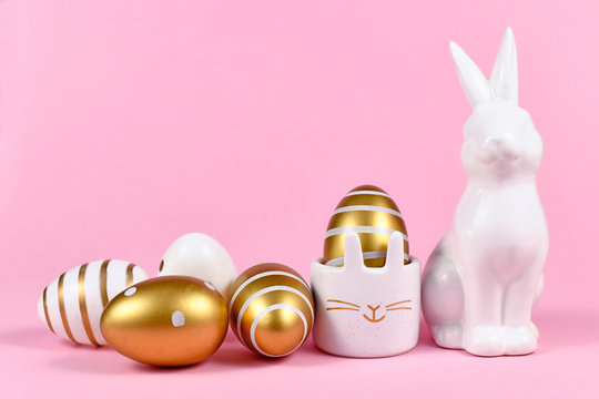 Golden and white painted easter eggs with dots and stripes, cute easter egg cup in shape of bunny and rabbit figure on pink background