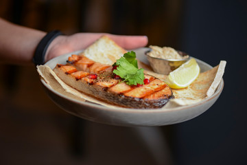 Waiter holds a plate of tasty food, Grilled salmon steak with lemon and pomegranate seeds on a white plate.