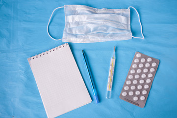 Notepad, pen, medical mask, tablets, thermometer on a blue background